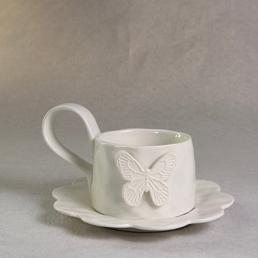 A white ceramic teacup with a matching saucer, featuring an embossed butterfly design on the front, set against a neutral gray background.