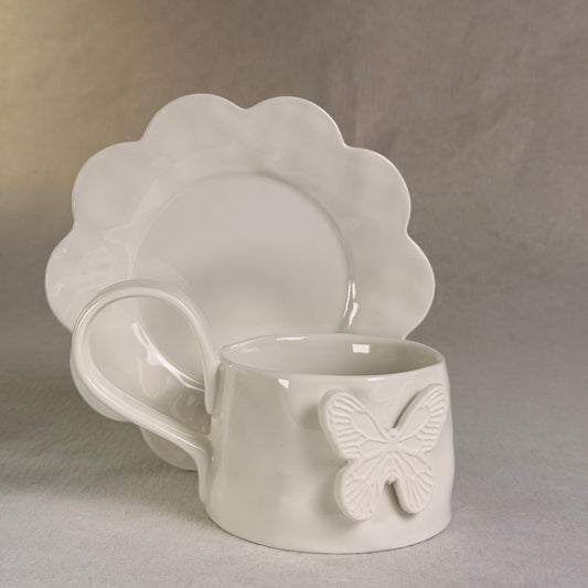 White ceramic cup and matching saucer set with a raised butterfly motif on the cup, photographed against a soft neutral fabric backdrop.