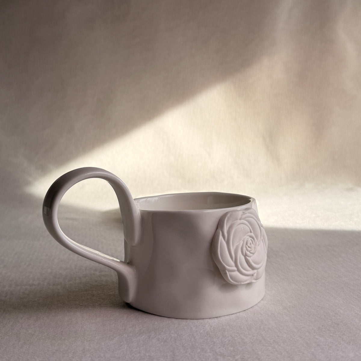 A matte white ceramic mug with a large handle and a raised rose decoration on the side, set against a draped fabric backdrop with soft light and shadow.