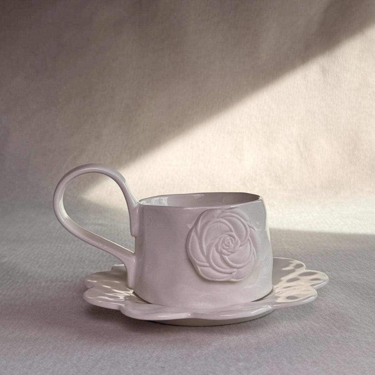 A white ceramic teacup with a matching saucer, featuring a raised embossed rose design on the side, set against a textured neutral background with dramatic diagonal lighting.