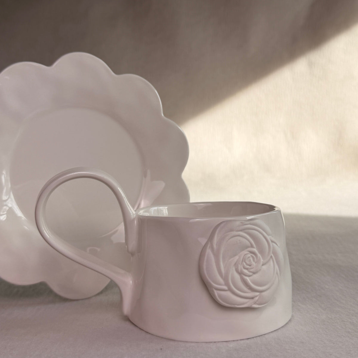 A white ceramic mug with a sculpted rose detail on the side, shown next to a matching scalloped saucer against a draped fabric backdrop with soft diagonal light.