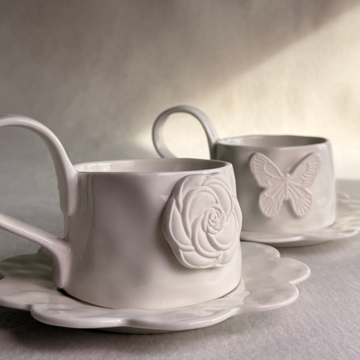 Two white ceramic teacups with matching saucers, featuring embossed floral and butterfly designs, set against a soft gray background with warm light shadows.