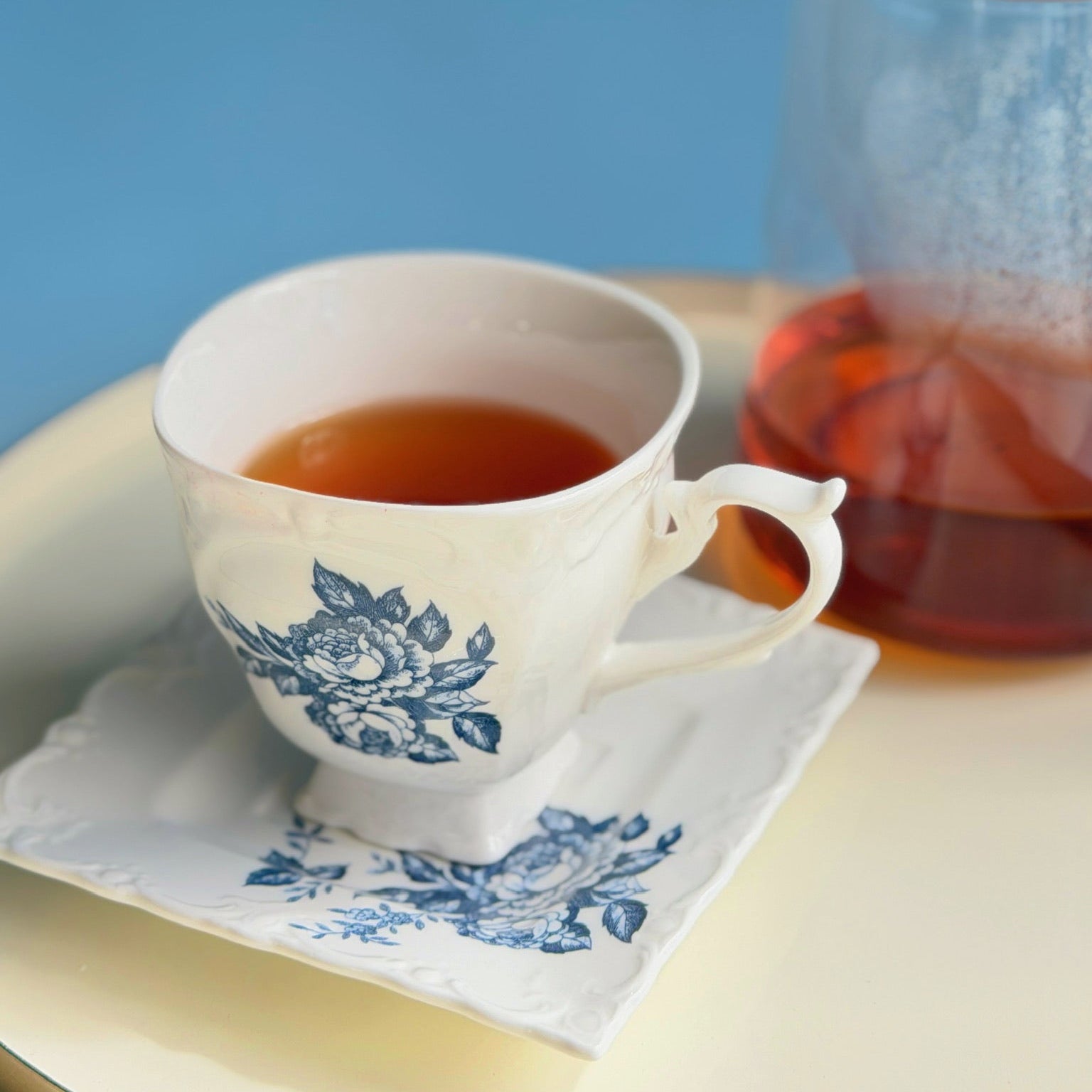Tea cup with floral design on a white napkin, next to a glass teapot on a blue background