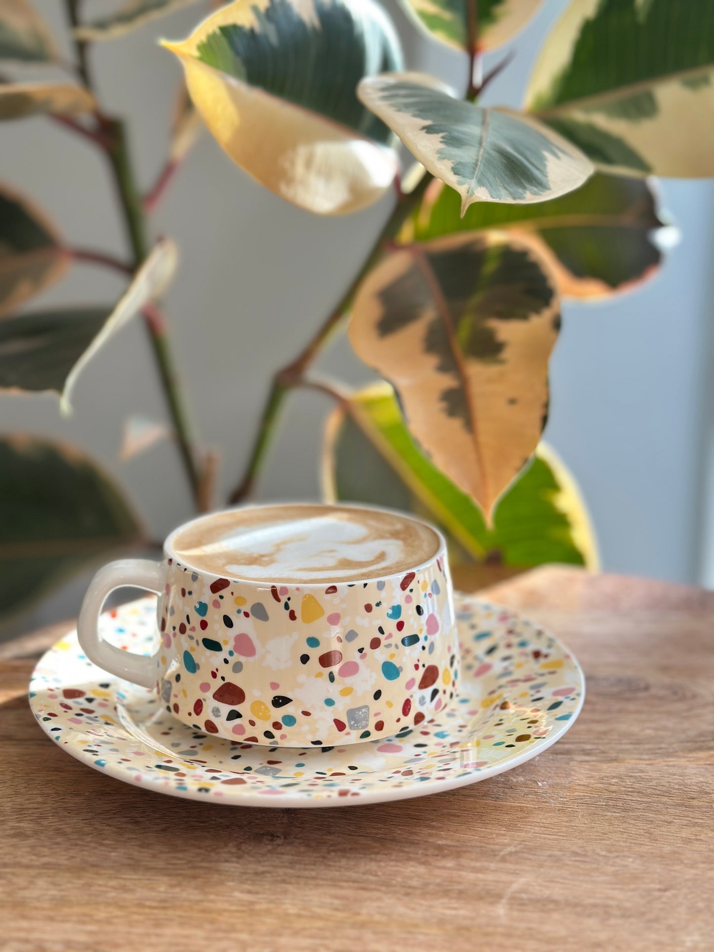 Colorful terrazzo-patterned tea cup and saucer on a wooden table with a plant in the background