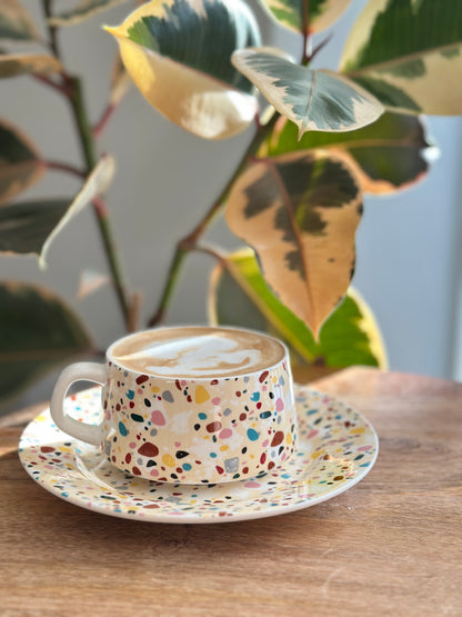 Colorful terrazzo-patterned tea cup and saucer on a wooden table with a plant in the background
