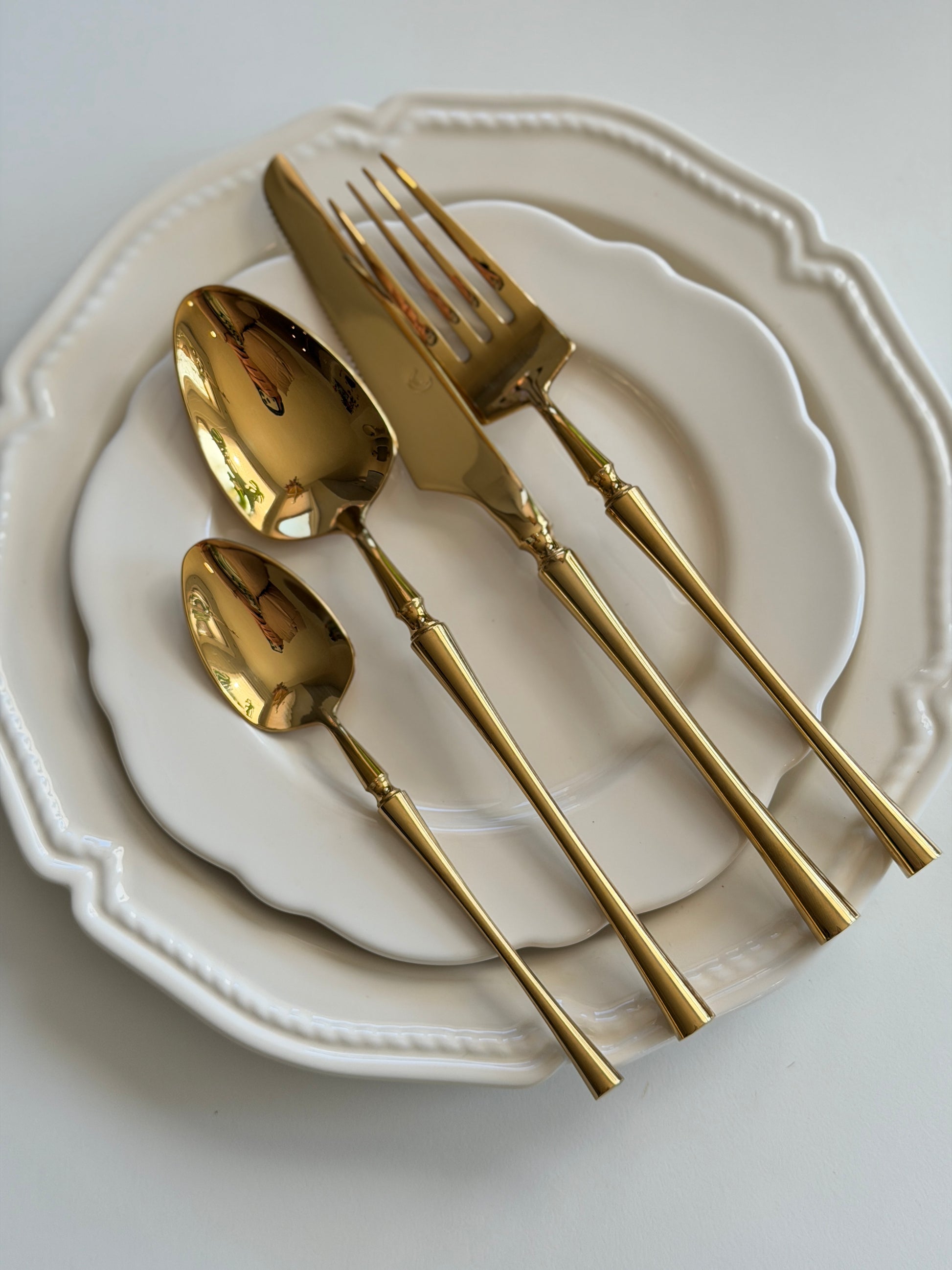 Gold polished fork, knife and two spoons with slim fluted handles, arranged on a scalloped white plate.