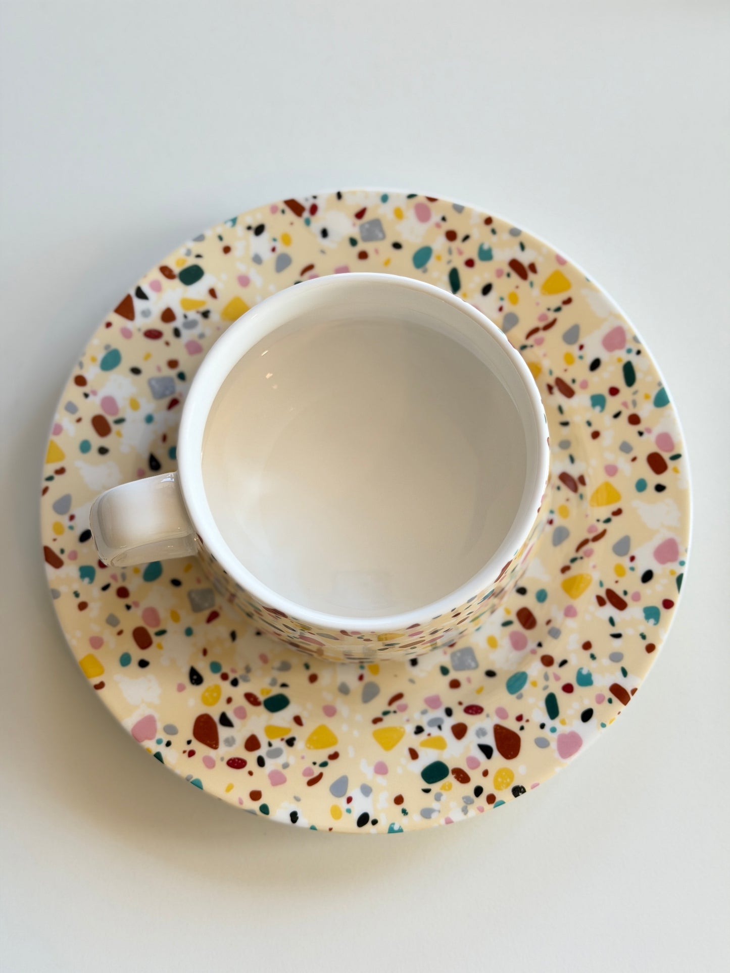 White ceramic tea cup and saucer with multicolored speckled pattern on a light gray background