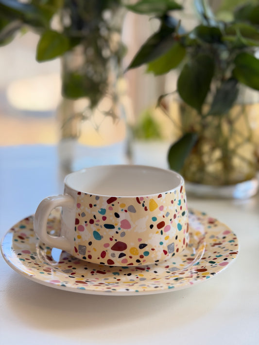 Colorful terrazzo-patterned tea cup and saucer on a white surface with blurred greenery in the background
