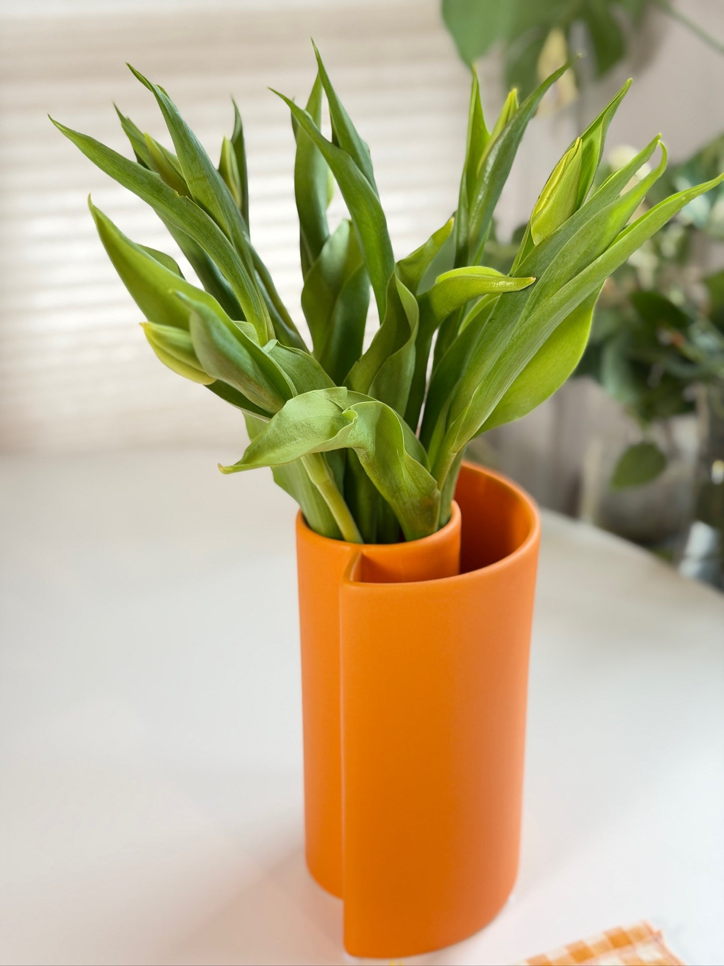 Orange Petal Ceramic Vase with overlapping wrapped form holding green tulip foliage on a white table.