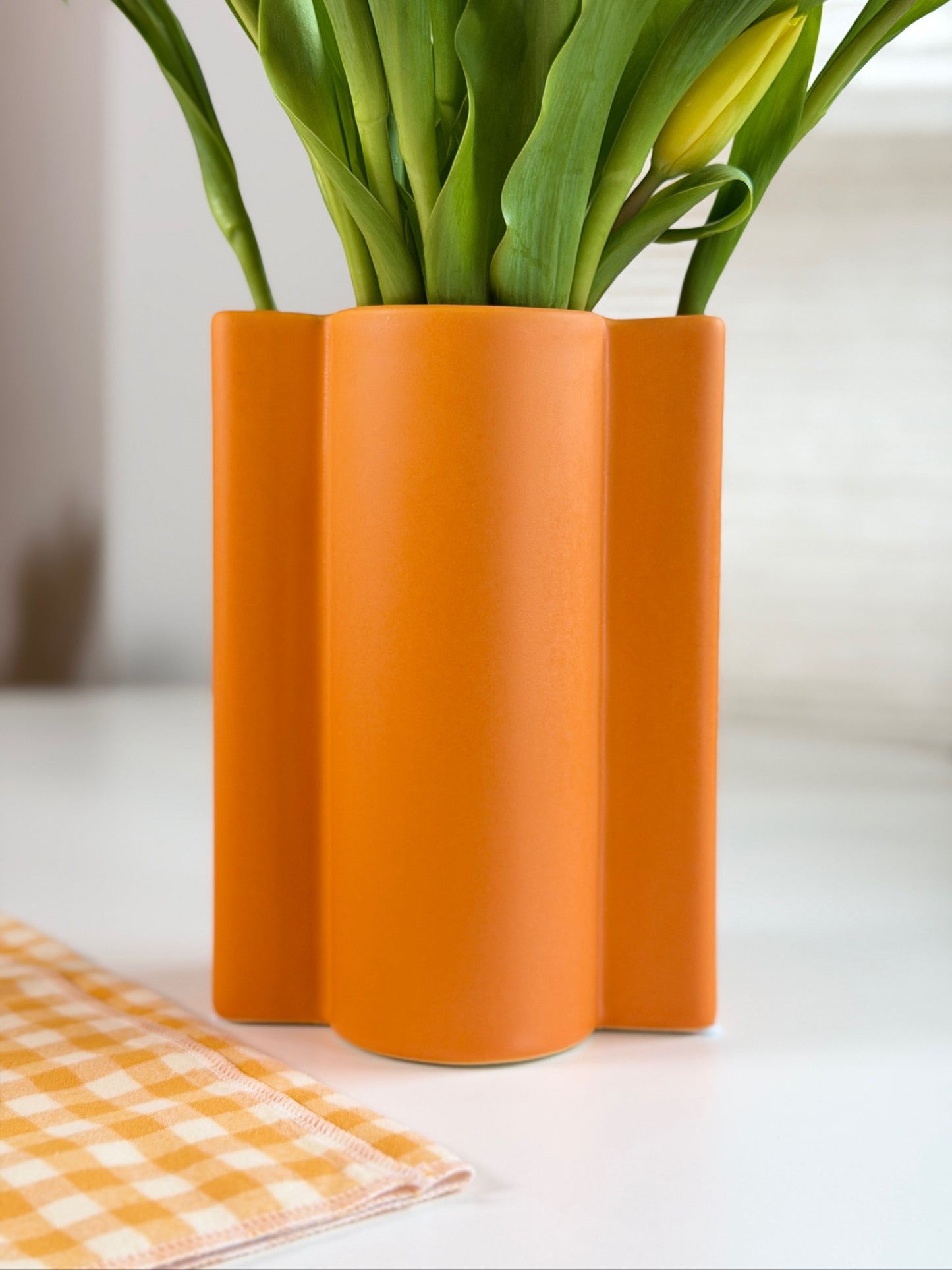 Orange wavy ceramic vase with green tulips on a white surface with an orange checkered cloth.