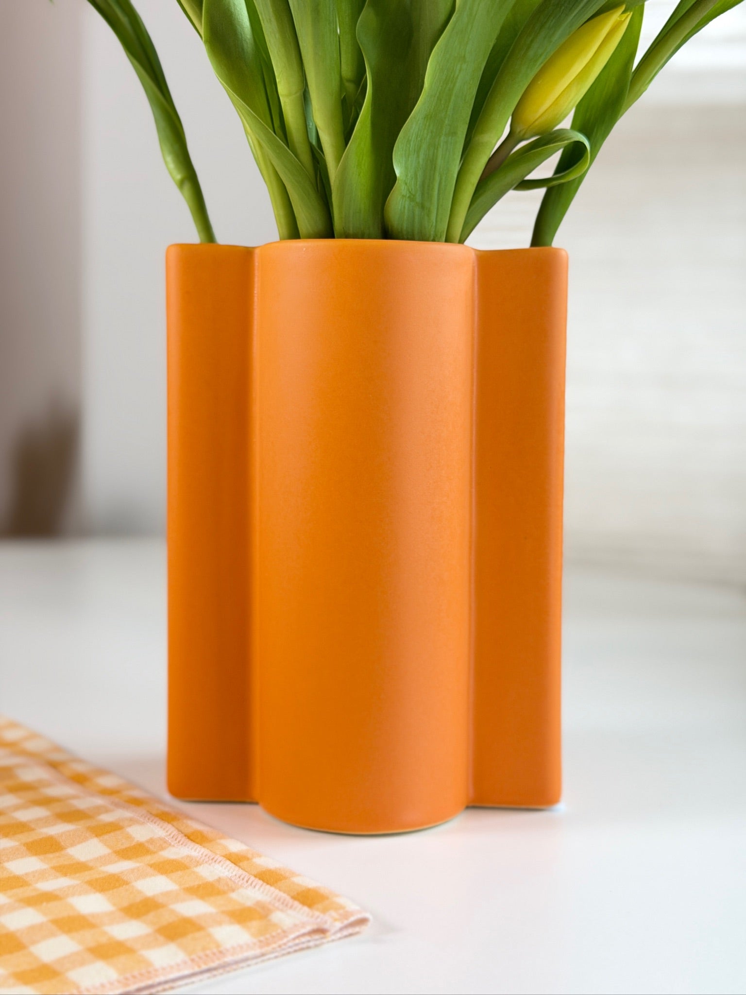 Orange wavy ceramic vase with green tulips on a white surface with an orange checkered cloth.