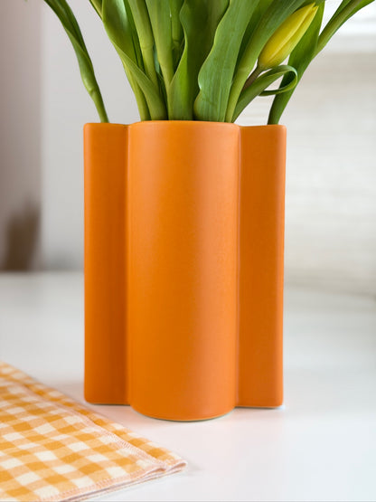Orange wavy ceramic vase with green tulips on a white surface with an orange checkered cloth.