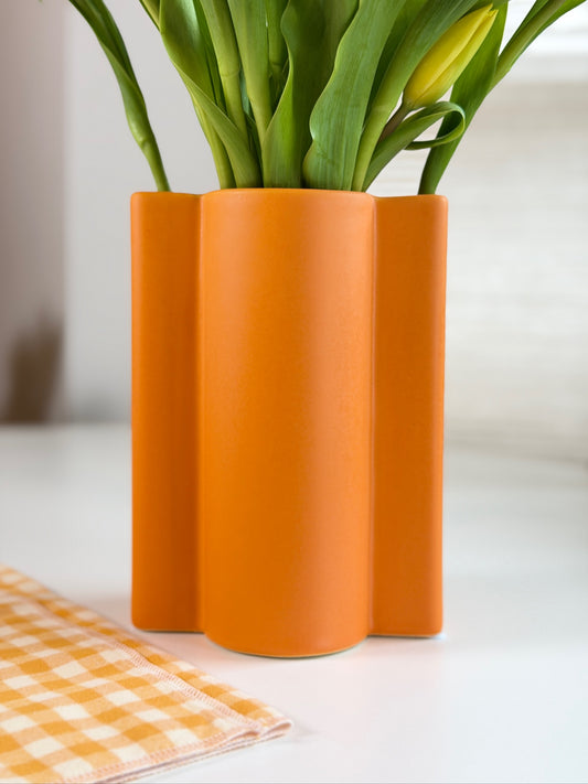 Orange wavy ceramic vase with green tulips on a white surface with an orange checkered cloth.