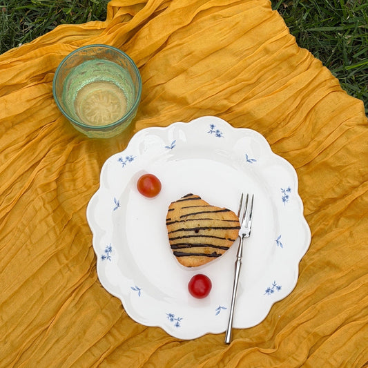 White porcelain blue floral patterned plate with pastry, tomatoes, and a glass on a yellow cloth on grass