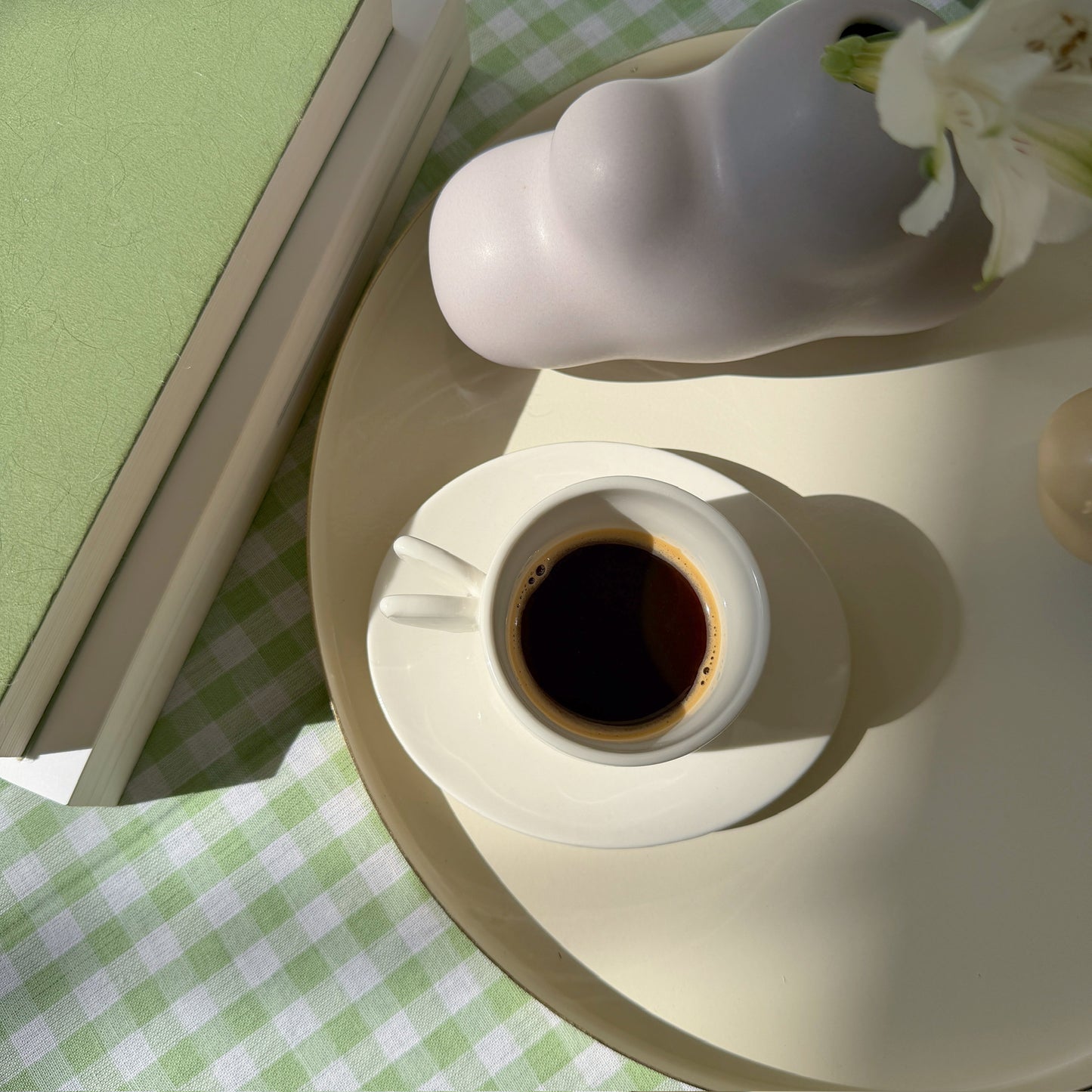 White cup of coffee on a round white table with a green checkered tablecloth.