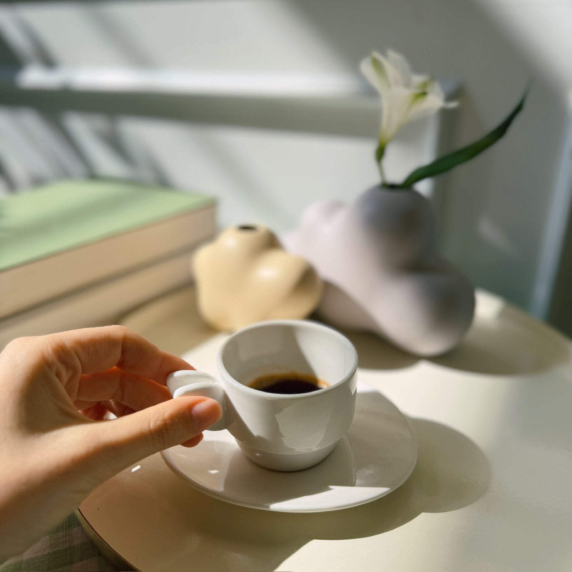 Hand holding a white coffee cup on a saucer with a vase and book in the background.