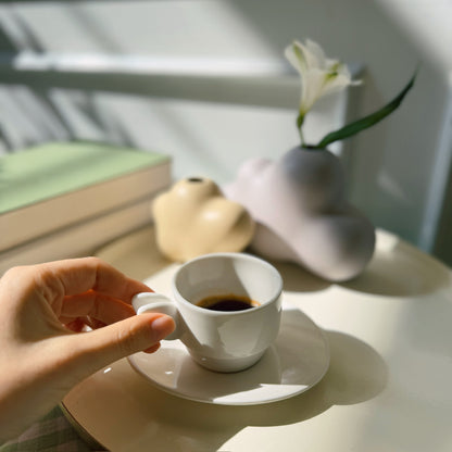 Hand holding a white coffee cup on a saucer with a vase and book in the background.