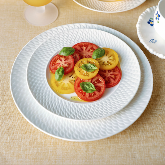 White textured ceramic dinnerware set on a beige tablecloth, featuring a large charger plate, a dinner plate, and a bowl holding sliced red and yellow tomatoes with fresh basil leaves. A yellow goblet, lemons, and a blue hydrangea are arranged around it.
