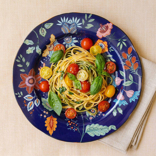 A top-down view of a beautifully decorated ceramic plate filled with spaghetti, cherry tomatoes, and fresh basil, set on a beige linen tablecloth with a folded napkin.