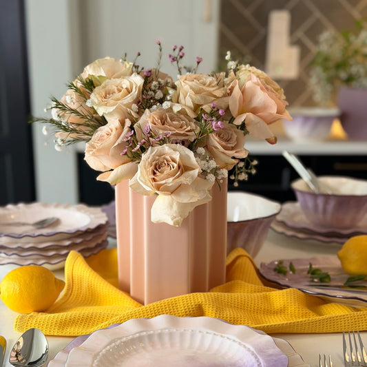 Blush pink ridged bookcase ceramic vase holding roses and baby's breath as a centerpiece on a lilac plate table setting.