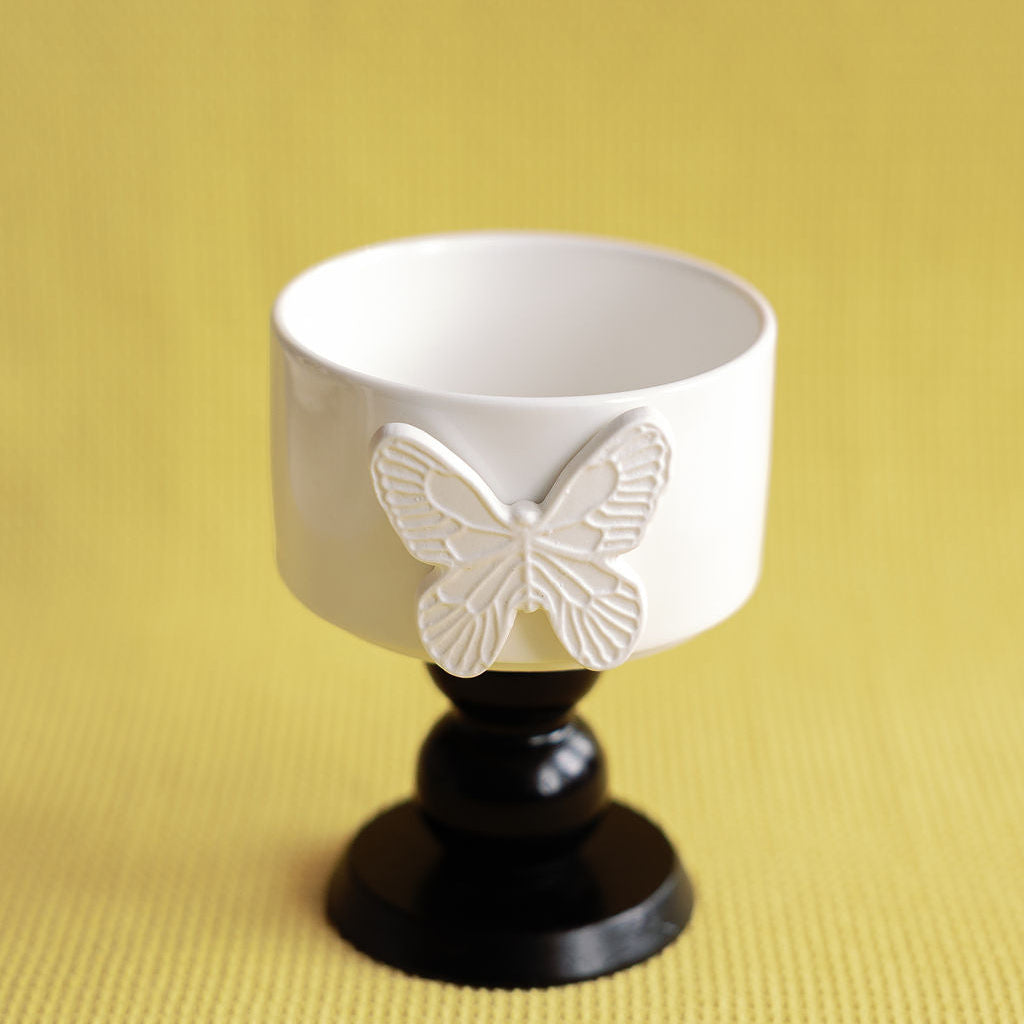 White ceramic cup with butterfly design on a black pedestal against a yellow background