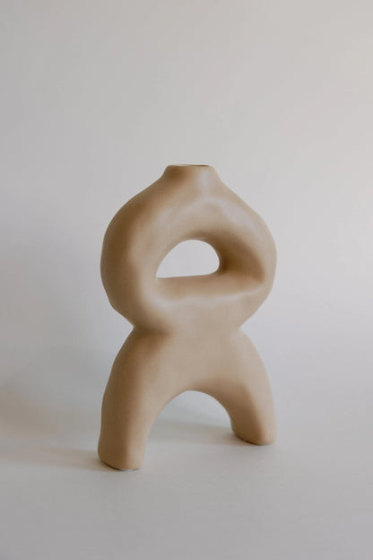 Beige sculptural bellow bud vase with looped body, arched legs and small top opening, matte ceramic front view