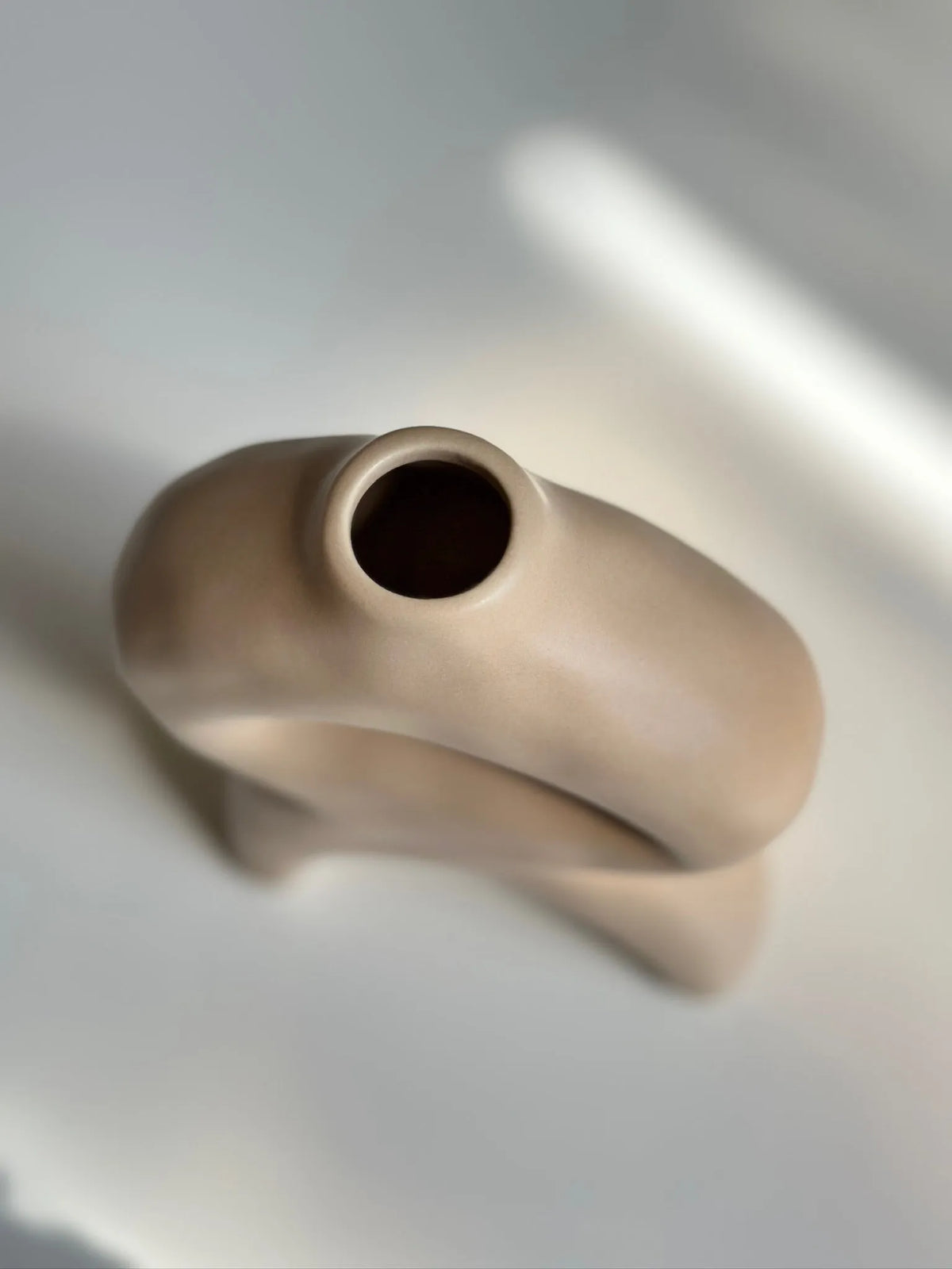 Tan bellow bud vase top-down close-up showing the round opening and curved looped body against a soft-lit surface