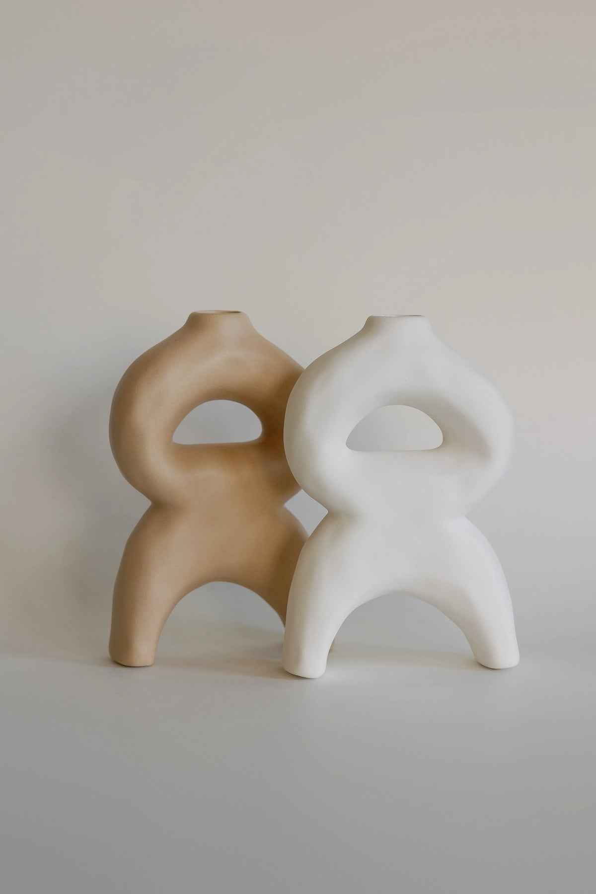 Tan and white sculptural bellow bud vases side by side, looped figure-eight bodies on arched legs, matte ceramic