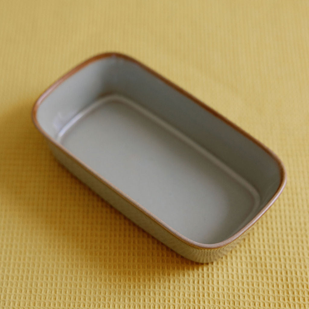 Ceramic glazed serving dish in grey-green with a rust rim, angled on a yellow waffle-weave surface.