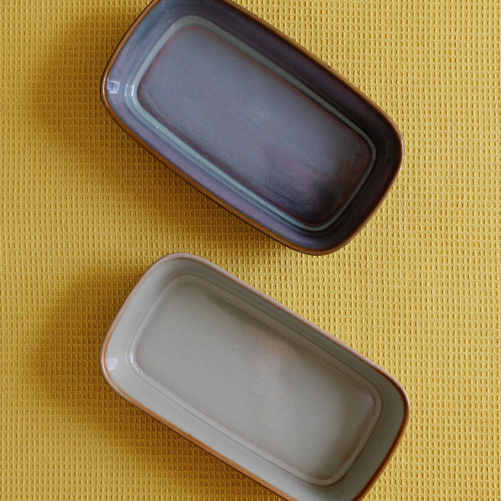Two ceramic glazed serving dishes overhead, one purple-blue and one tan-grey, on a yellow textured cloth.