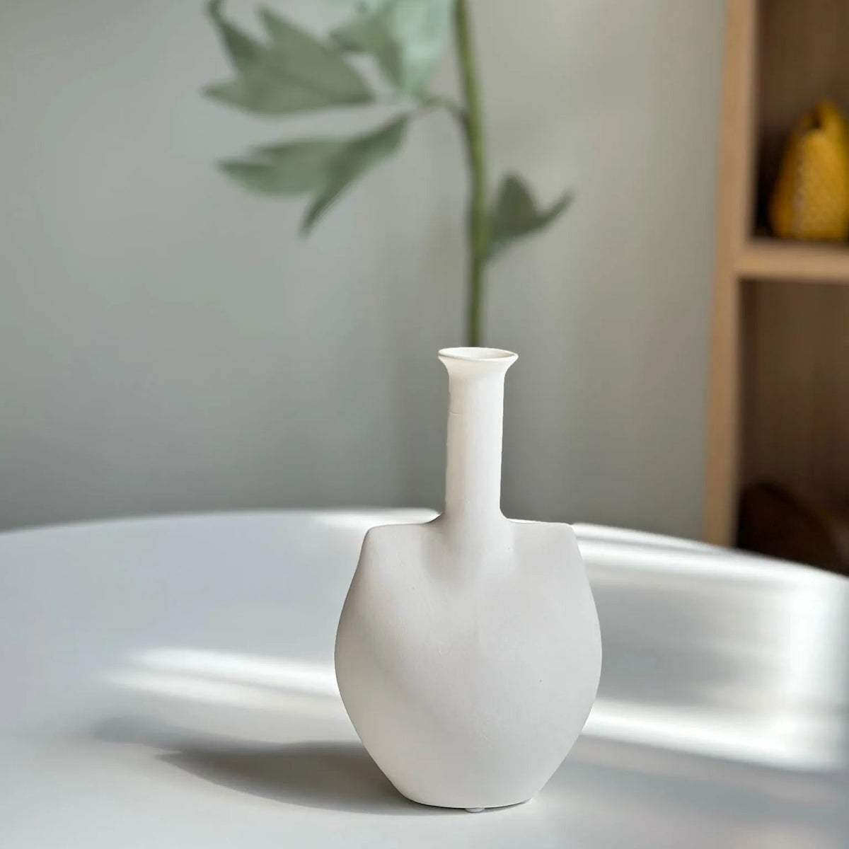 White ceramic trowel bud vase styled on a table with a green leafy stem behind it