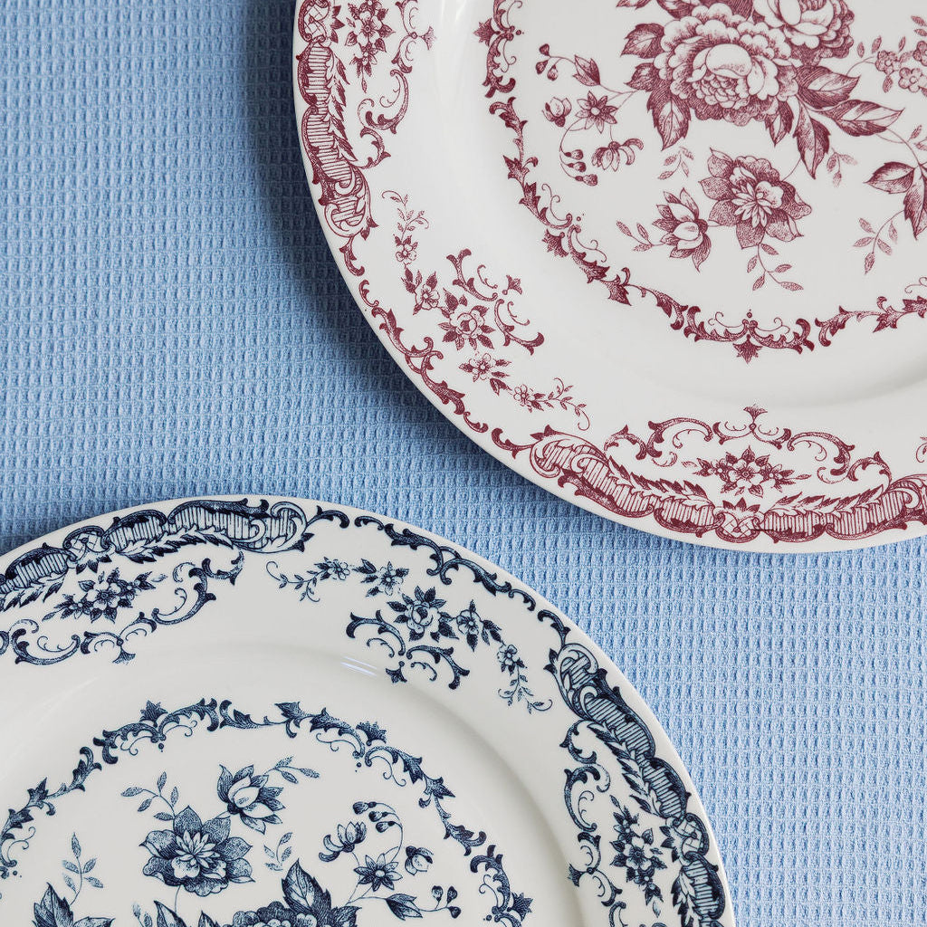 Two Chantilly Lace porcelain plates, one red floral and one blue floral, side by side on a pale blue surface.