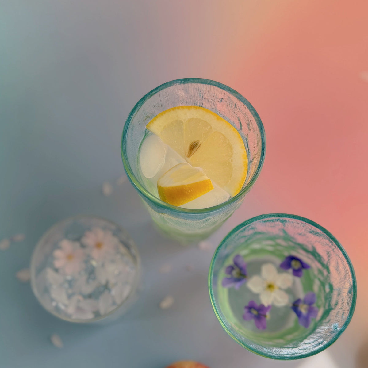 Coastal Hues glasses from above with lemon, ice and floating violets, styled on a pastel ombre surface.