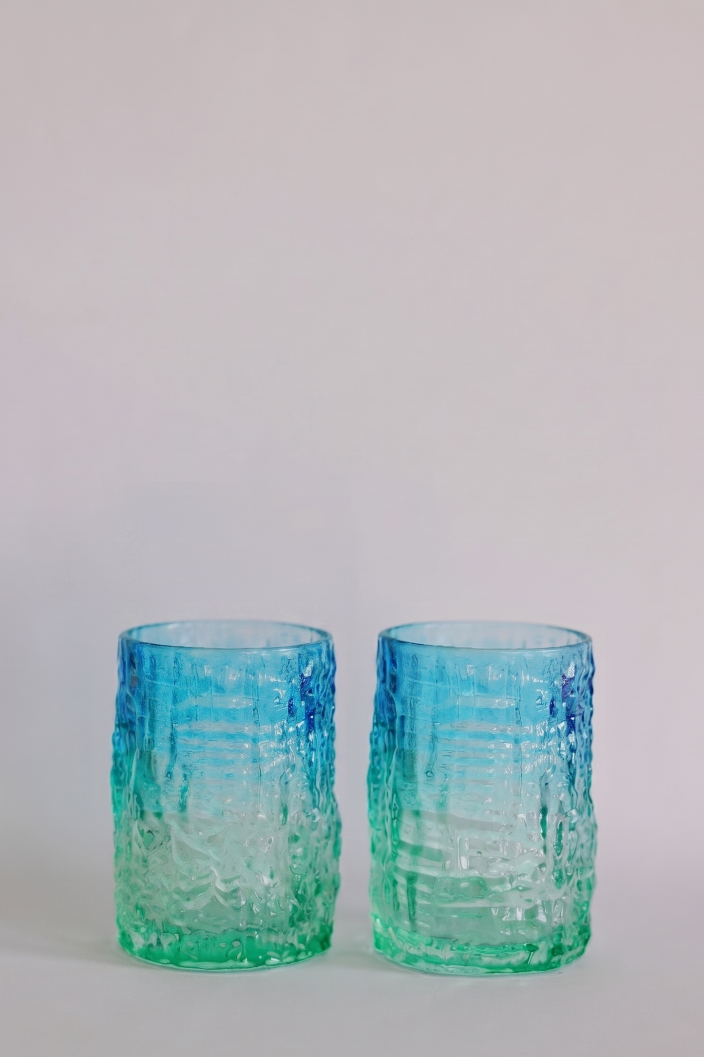 Two textured glass tumblers with a blue-to-green gradient, standing side-by-side on a light gray surface against a neutral light gray background.