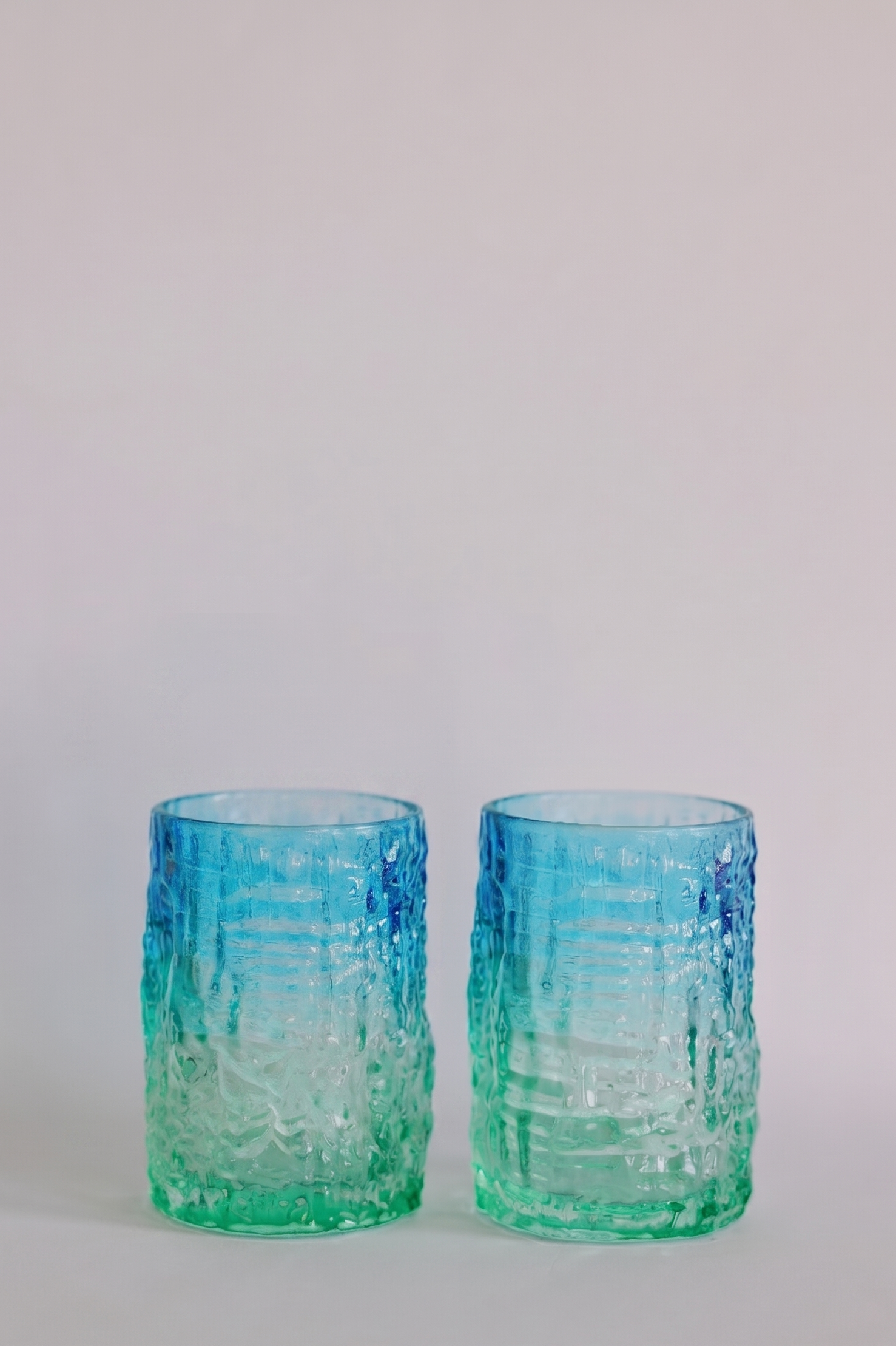 Two textured glass tumblers with a blue-to-green gradient, standing side-by-side on a light gray surface against a neutral light gray background.