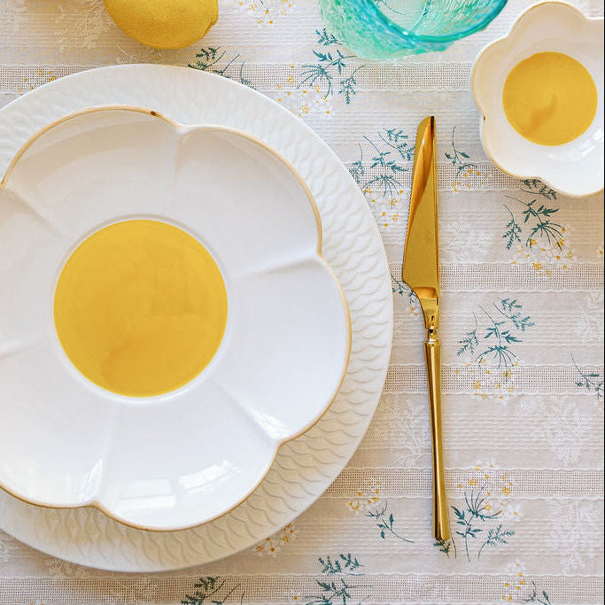 A set table with white flower-shaped plates, yellow centers, and blue glassware, surrounded by lemons and floral tablecloth.