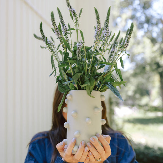 Person holding the white dotted Dotty Cylinder Ceramic Vase with purple veronica blooms, outdoor scene.