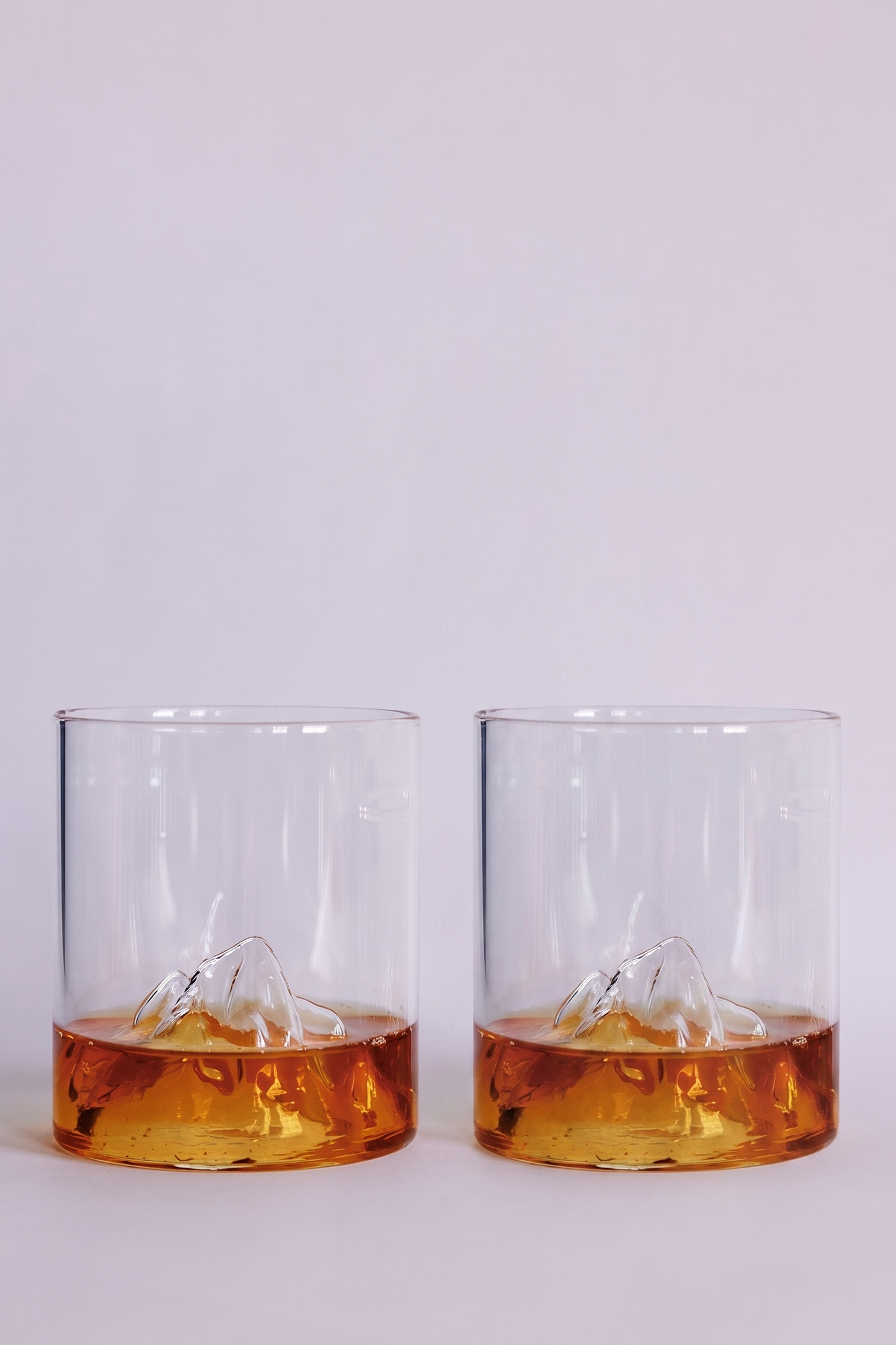 Two clear rocks glasses with a sculpted mountain design at the base, shown on a clean white background with amber-colored liquid inside.