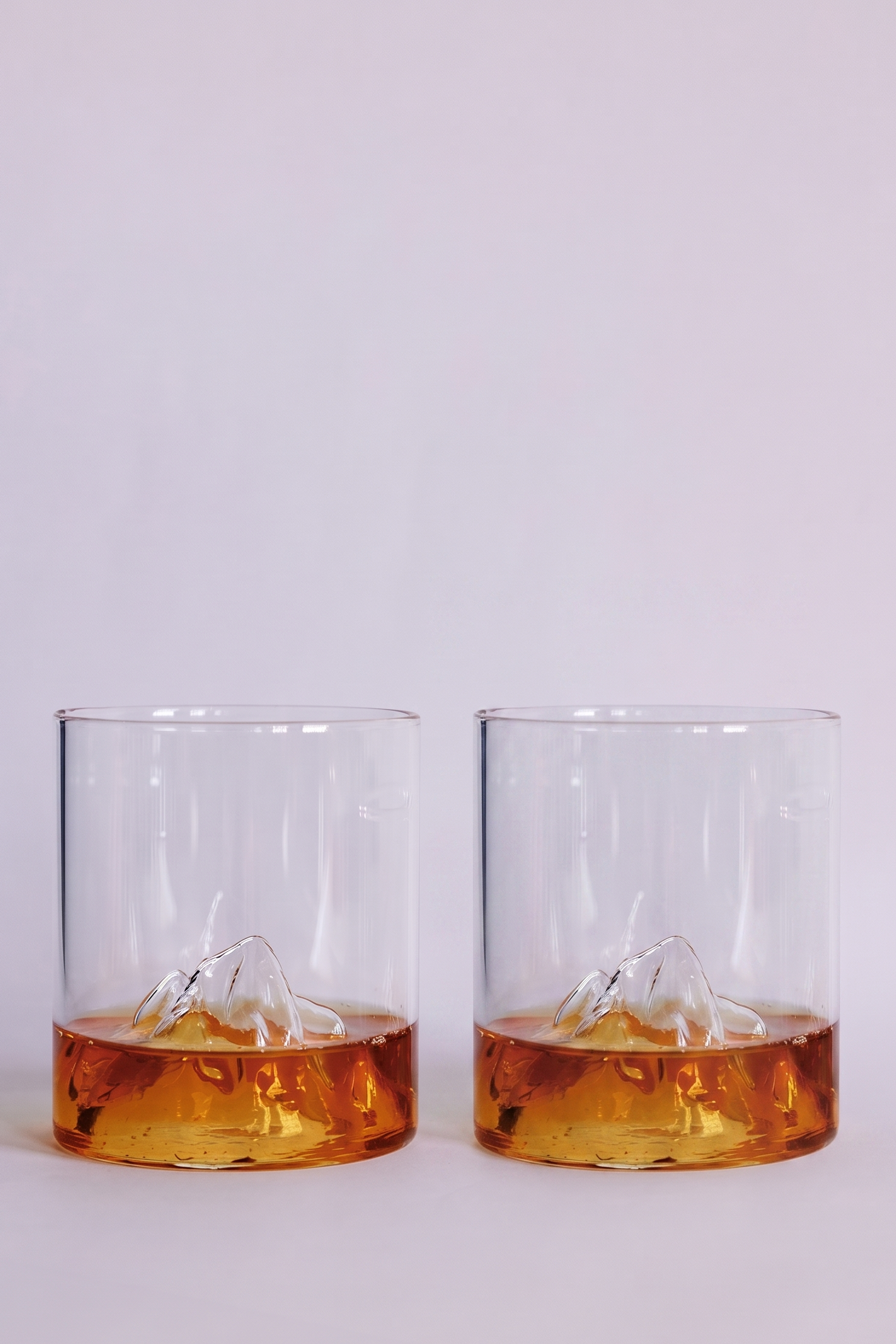 Two clear rocks glasses with a sculpted mountain design at the base, shown on a clean white background with amber-colored liquid inside.