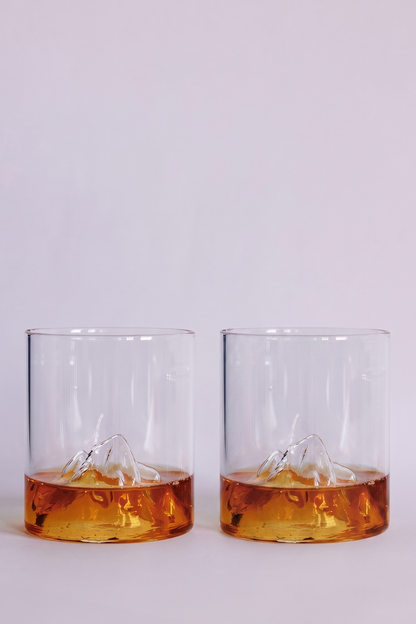 Two clear rocks glasses with a sculpted mountain design at the base, shown on a clean white background with amber-colored liquid inside.