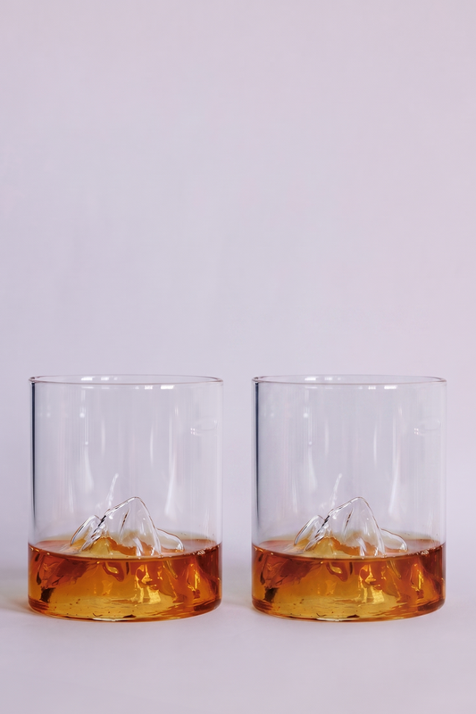 Two clear rocks glasses with a sculpted mountain design at the base, shown on a clean white background with amber-colored liquid inside.