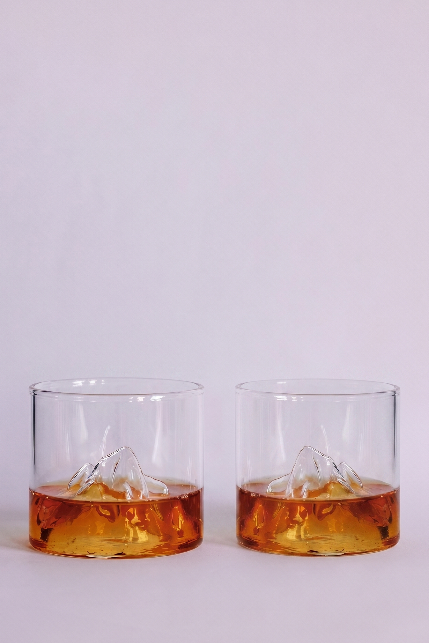 Two clear rocks glasses with a mountain design inside, partially filled with amber-colored whiskey, set against a plain light gray background.