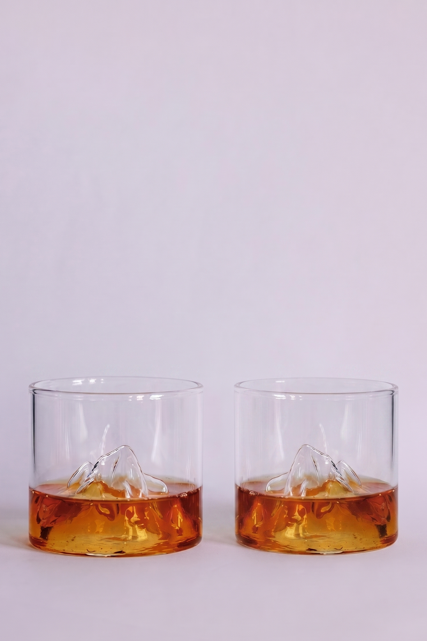 Two clear rocks glasses with a mountain design inside, partially filled with amber-colored whiskey, set against a plain light gray background.