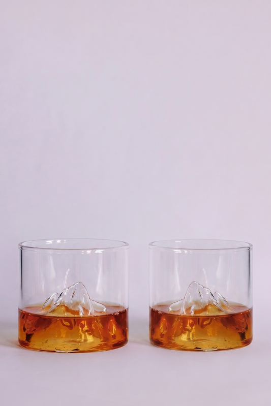Two clear rocks glasses with a mountain design inside, partially filled with amber-colored whiskey, set against a plain light gray background.
