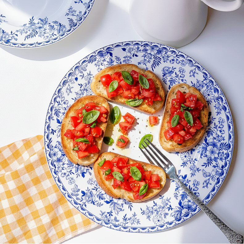 A blue and white floral ceramic plate holding four slices of toasted bread topped with tomato and basil bruschetta, styled on a white surface with a white pitcher, another plate, and a yellow-and-white checkered napkin.