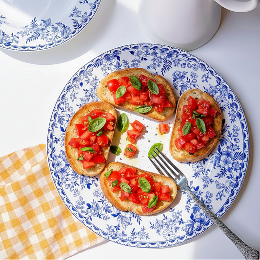 A blue and white floral ceramic plate holding four slices of toasted bread topped with tomato and basil bruschetta, styled on a white surface with a white pitcher, another plate, and a yellow-and-white checkered napkin.