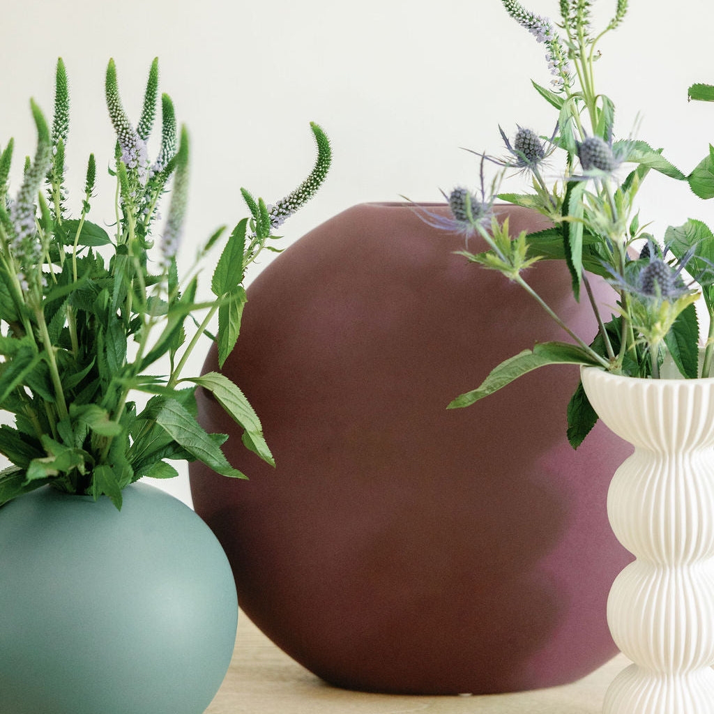 Luna Ceramic Vase in plum, flat round vase styled on a wood table beside green and white vases with greenery.