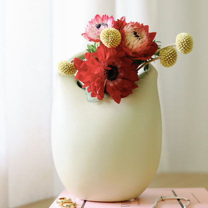 Cream ceramic basket bud vase filled with red and pink flowers and yellow billy buttons, on stacked books.