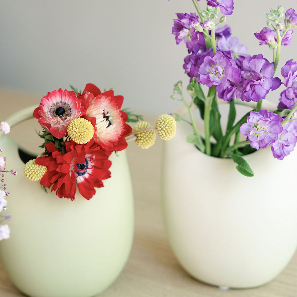 Two green ceramic basket bud vases holding red anemones and purple stock flowers on a wood table.
