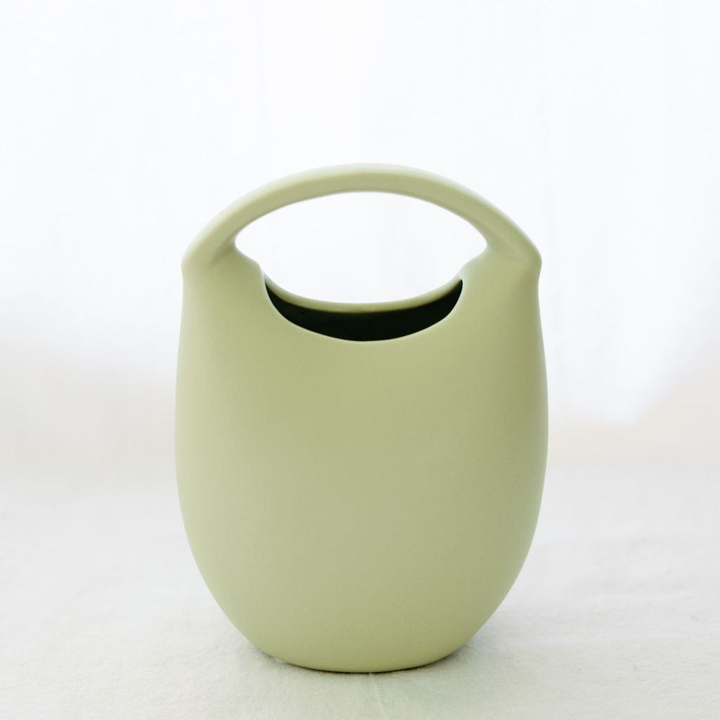 Matte sage green ceramic basket bud vase with arched top handle, shown empty on a white backdrop.